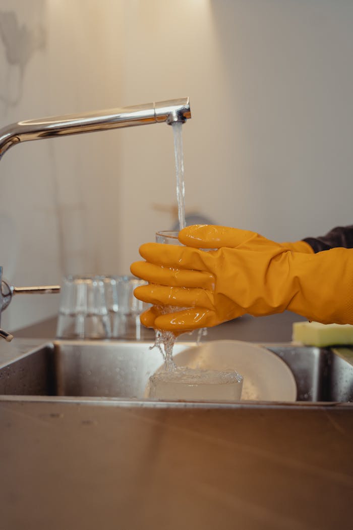 Close-up of hands in yellow gloves washing dishes under a faucet with flowing water.